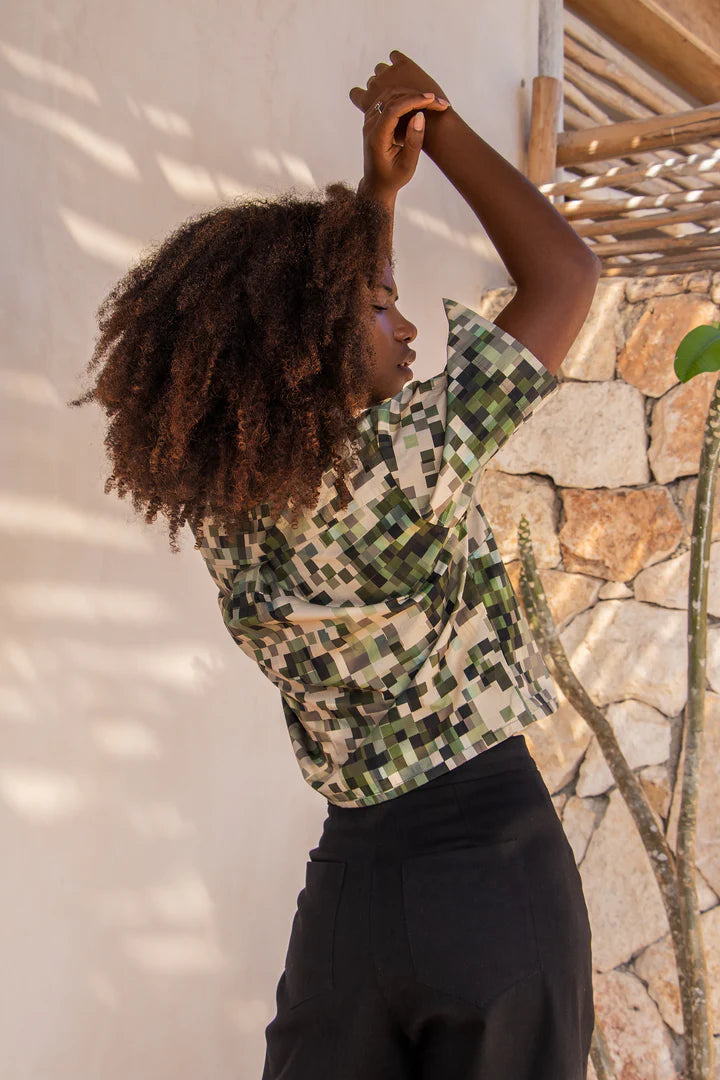 Back view of a woman wearing the Almeria Top by MAS in Camo, a slightly cropped button up short sleeved shirt with a large collar and a pixellated print in green and earth tones. She's wearing it with the Vienna Pants in Black, and standing outside by a stone wall. 