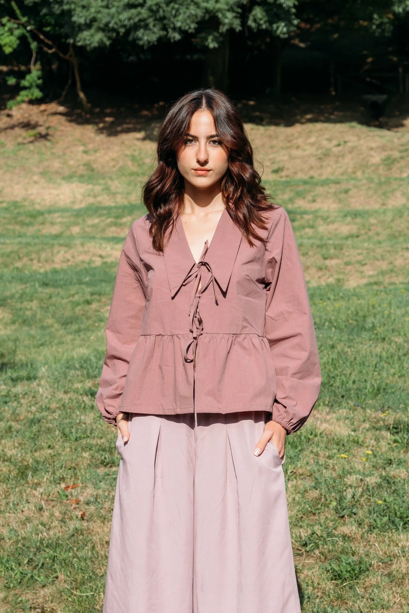A woman wearing the Siena Pants by MAS in Raspberry, wide legged trousers with an elastic waist. She is wearing them with a pink jacket and standing outdoors. 