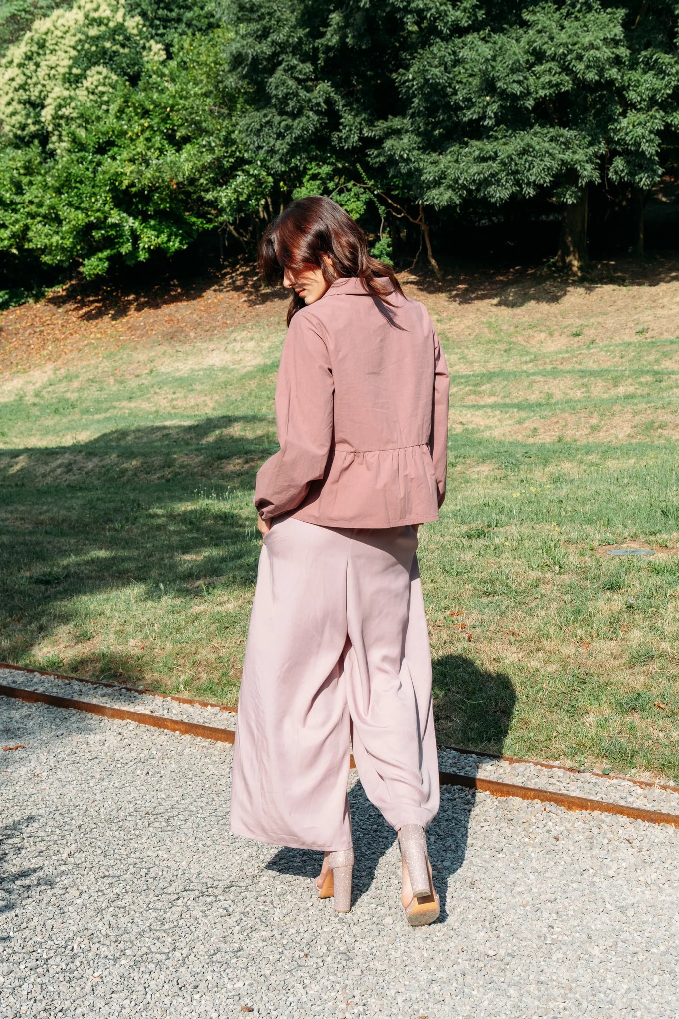 Back view of a woman wearing the Siena Pants by MAS in Raspberry, wide legged trousers with an elastic waist. She is wearing them with a pink jacket and standing outdoors. 