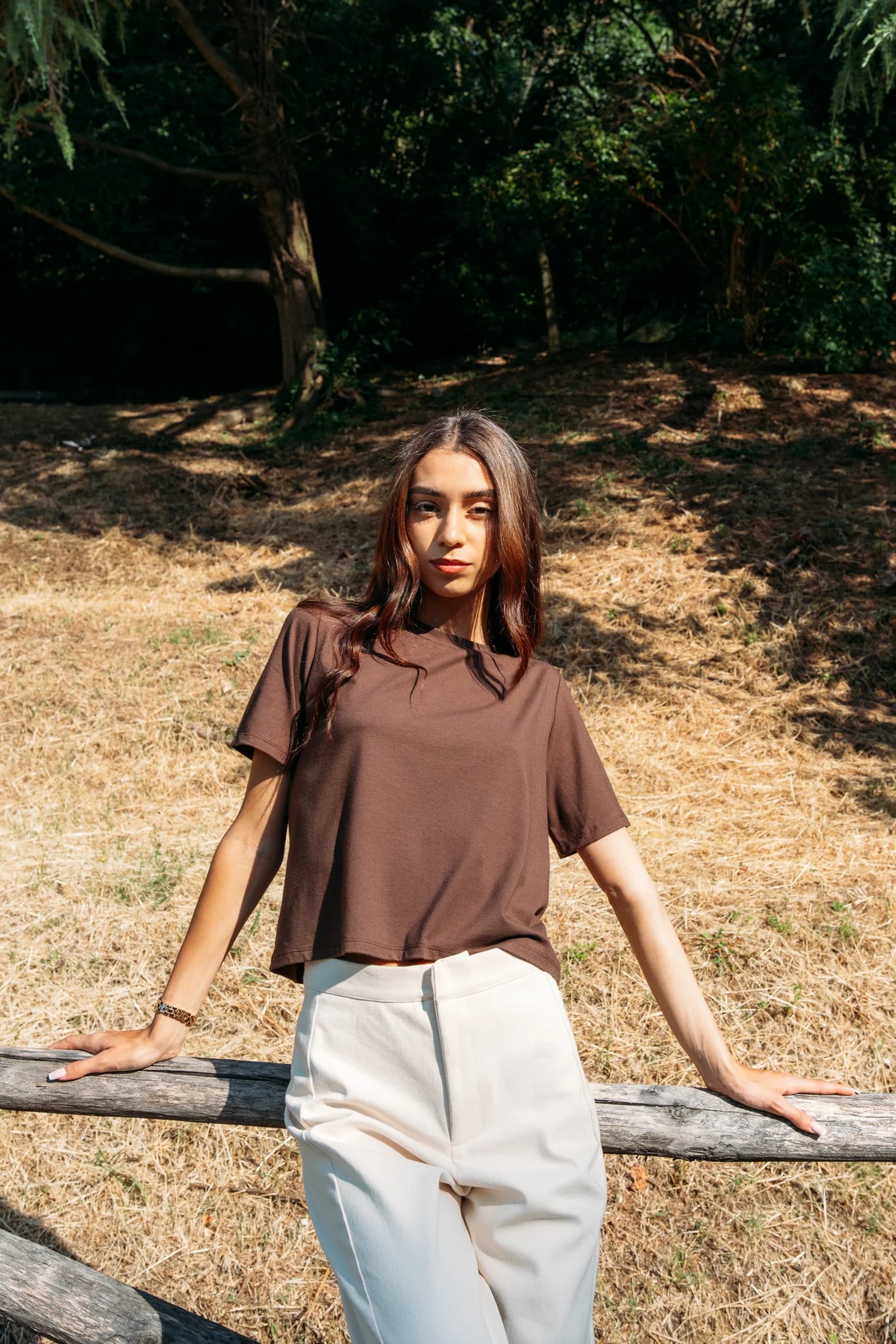 A woman wearing the Santiago T-Shirt by MAS in Cacao, a classic tee with a slightly cropped and boxy fit. She is wearing it with beige pants leaning on a log fence with a forest behind her. 
