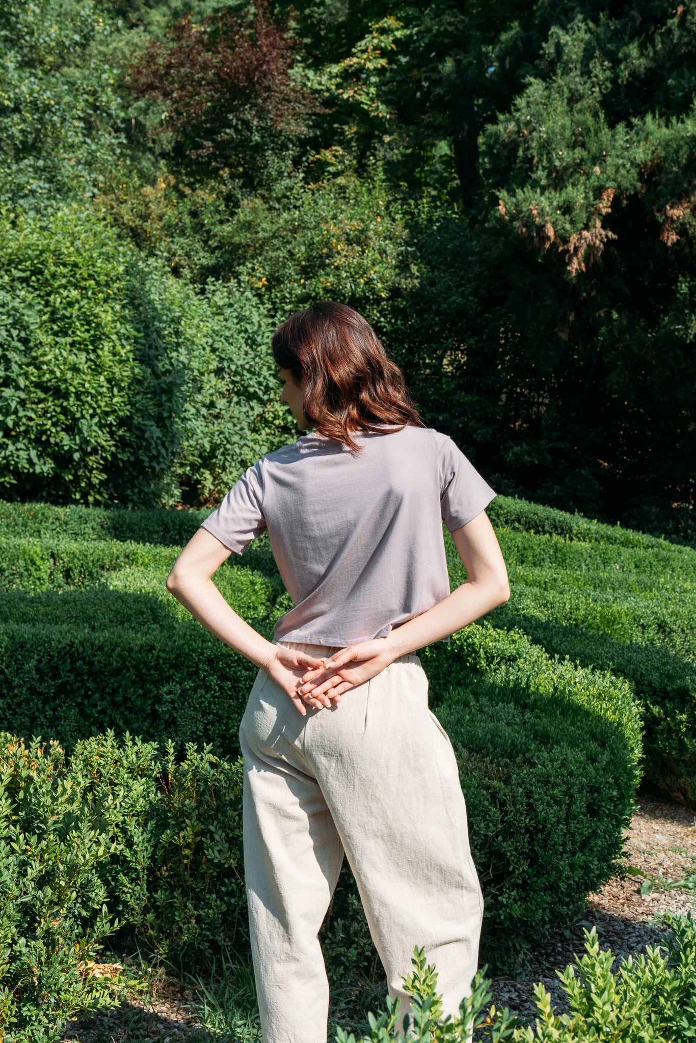 Back view of a woman wearing the Santiago T-Shirt by MAS in Lilac, a classic tee with a slightly cropped and boxy fit. She is wearing it with beige pants and standing outside with greenery behind her. 