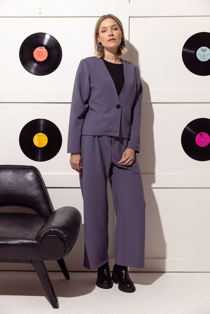 A woman wearing the Bowie Jacket by Cherry Bobin in Mauve featuring a V-neck, a single button closure, long sleeves and a short and slightly boxy shape. She is wearing it with matching pants and standing in front of a white wall with vinyl records hanging on it.