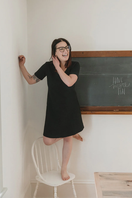 A woman wearing the Meadow Dress in Slub by Blondie Apparel in Black, standing on a white chair in front of a chalkboard