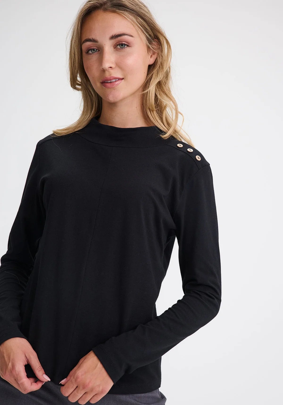 Waist up view of a woman wearing the Carleton Top by Message Factory in a Black, a long-sleeve top with button details on the shoulder. She is wearing it with grey bottoms and standing in front of a white background. 
