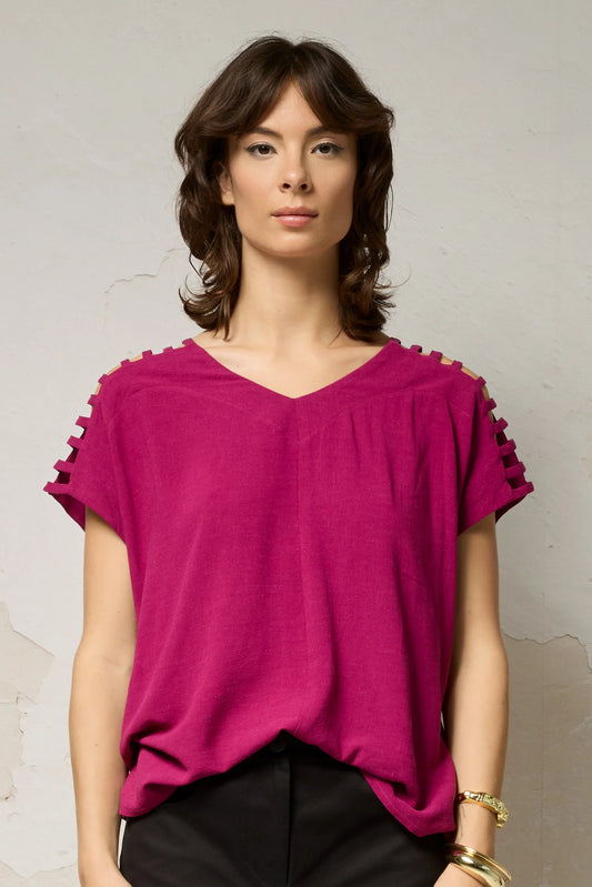 Hip-up view of a woman wearing the Mia Top by Cokluch in Dahlia pink, a short sleeved top with openwork ladder-like cutouts at the shoulders, a loose fit, and V-neck. She is wearing it with black pants and standing against a plain background. 