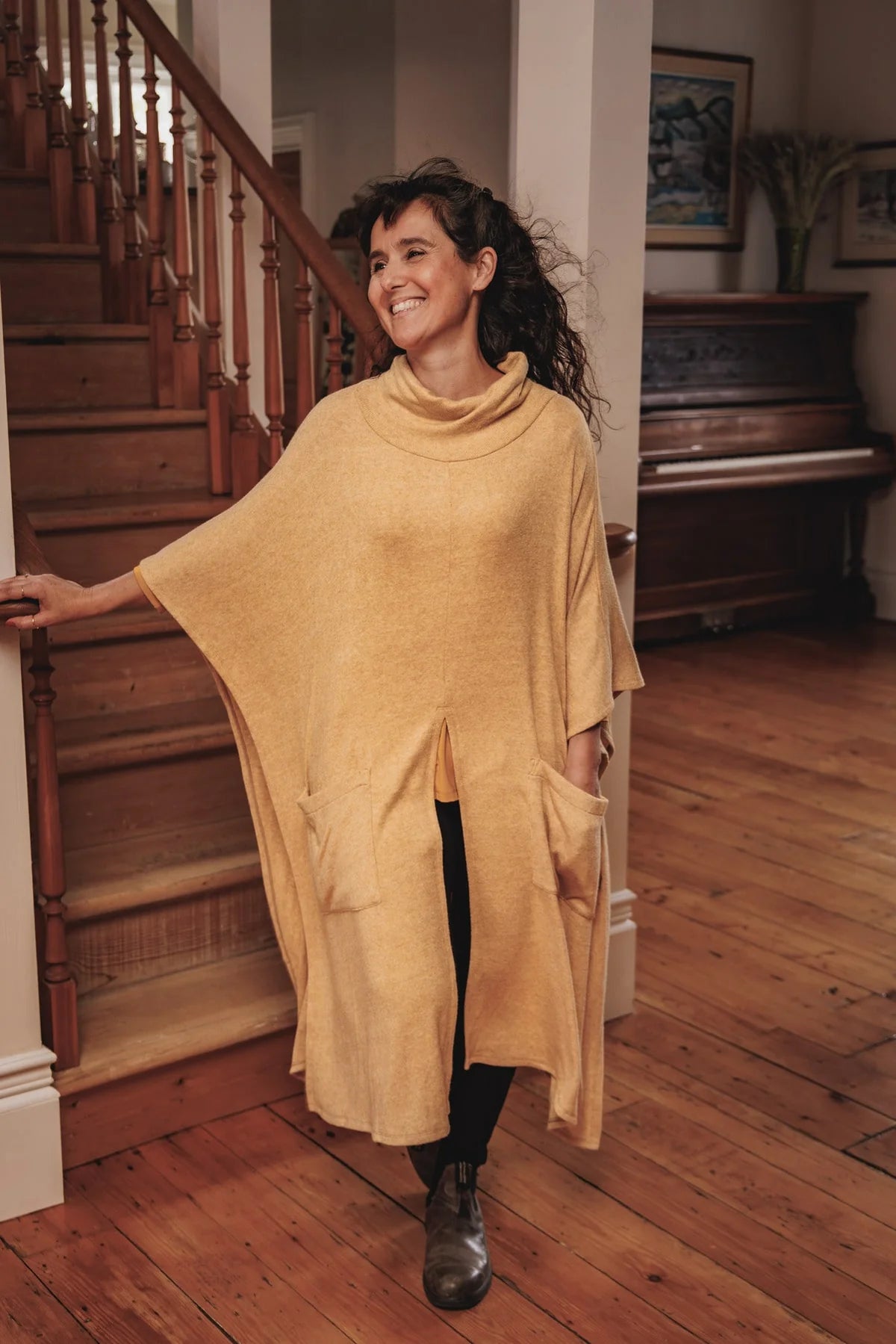 A woman wearing the Ora Poncho by MOOVment in Wheat, a mid-calf length poncho with a cowl neck, open sides, front slit, and large patch pockets. She is standing at the bottom of a wooden staircase.