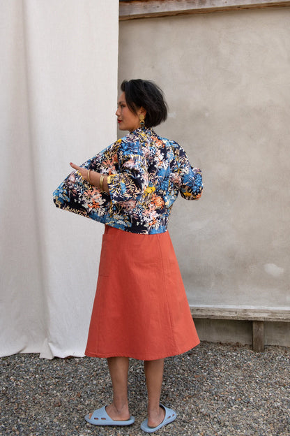 Back view of a woman wearing the Nymphea Screenprinted Skirt by Kazak in Red Pysanka with a floral jacket, she is standing outside in front of white curtain. 