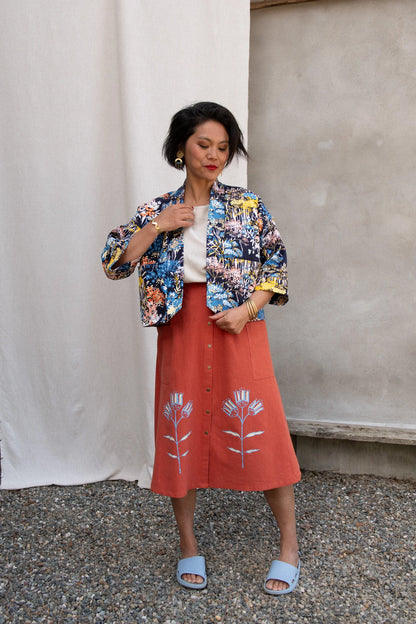 A woman wearing the Nymphea Screenprinted Skirt by Kazak in Red Pysanka with a floral jacket, she is standing outside in front of white curtain. 