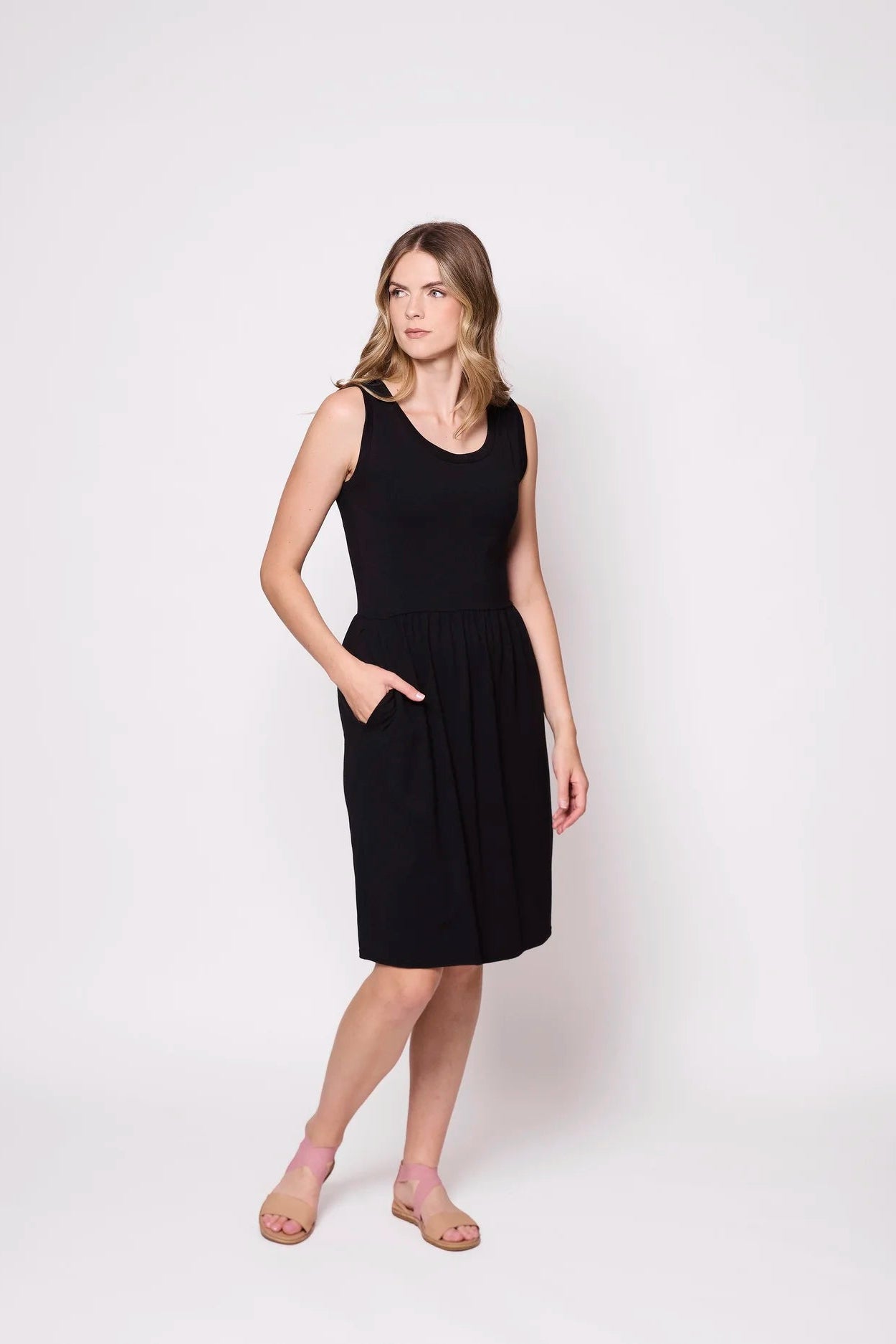 A woman wearing the Mai Dress by Tangente in Black, a sleeveless dress with a scooped neck and gathered knee-length skirt. She is standing against a white background