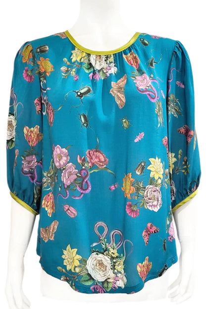 The Lydia Creeper Blouse by Mandala in Bayou is shown in front of a white background. It features a vintage floral print with snakes and insects, with a jewel neck, puffed 3/4 sleeves, and a scooped hem. 