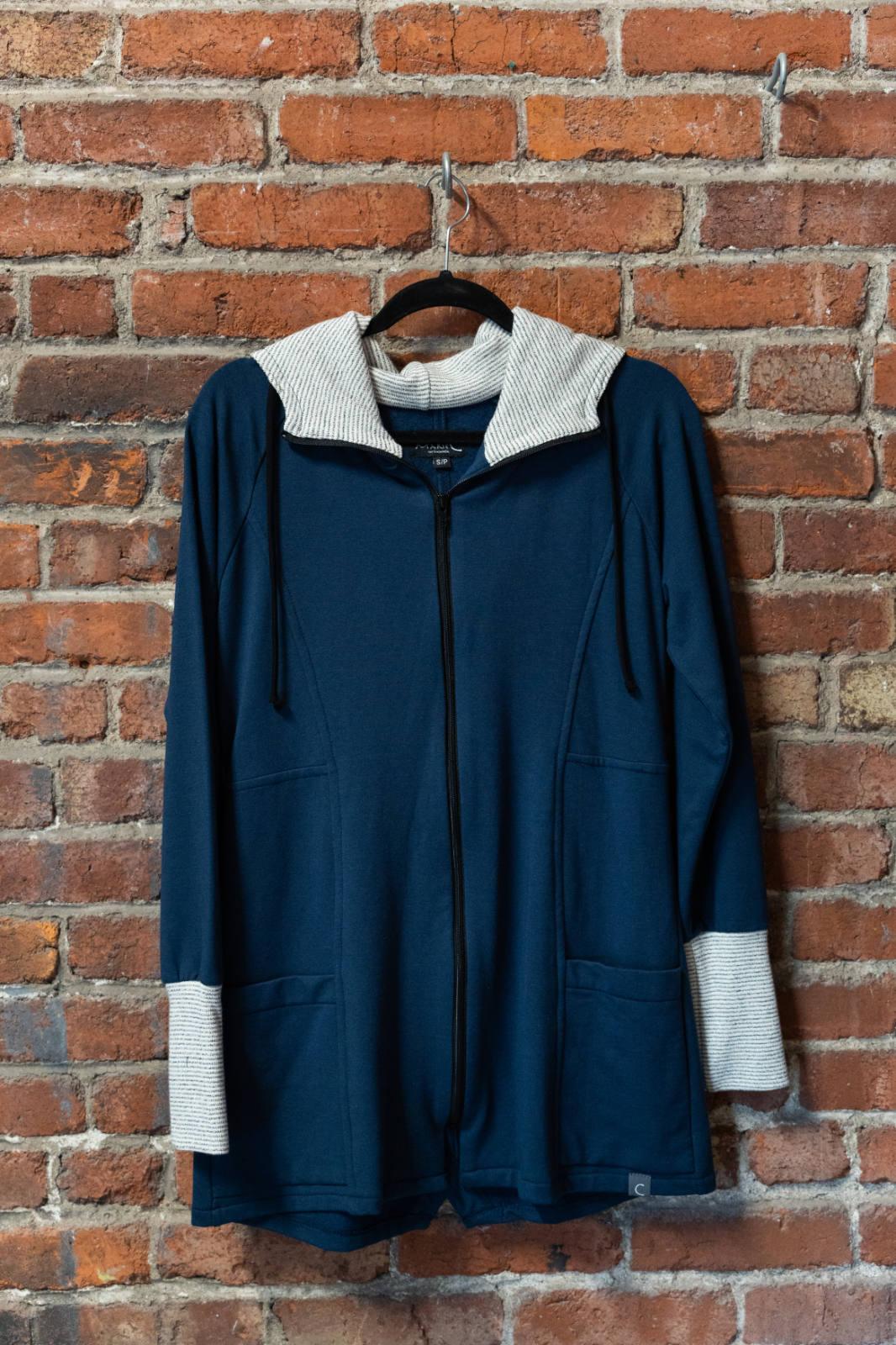 The Calma Jacket by Marie C in Navy, a mid-thigh length jacket with a hood and contrasting cuffs and lining, is shown hanging against a brick wall. 