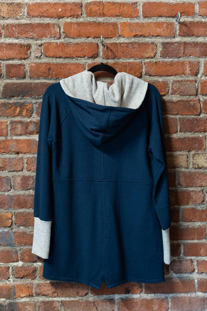 A back view of the Calma Jacket by Marie C in Navy, a mid-thigh length jacket with a hood and contrasting cuffs and lining, is shown hanging against a brick wall. 