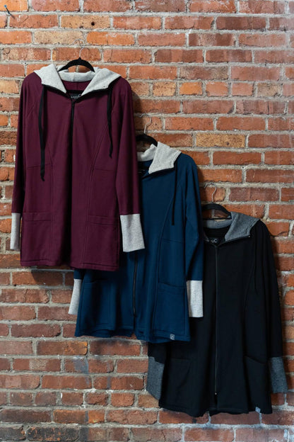 Three Calma Jackets by Marie C in Wine, Navy and Black,  a mid-thigh length jacket with a hood and contrasting cuffs and lining, are shown hanging against a brick wall. 