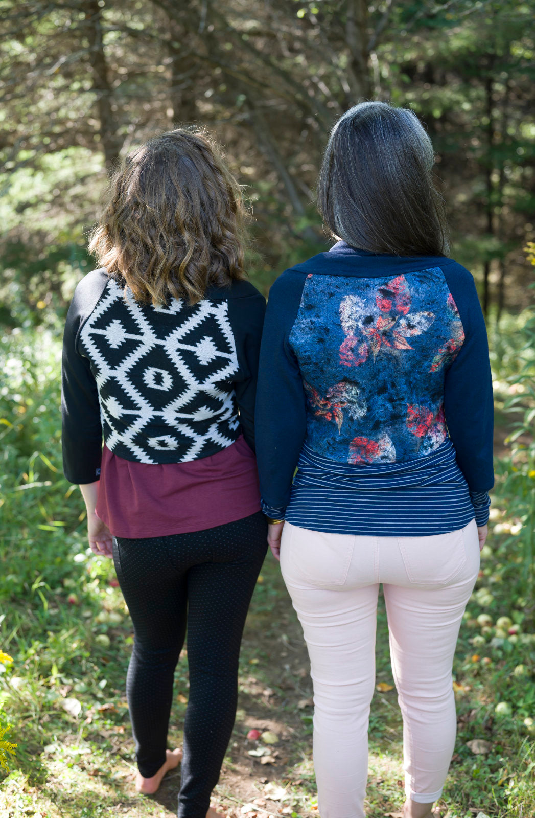 Back view of two women standing side to side in a forest, wearing the Boreal Bolero by Marie C. One is wearing the Black and one is wearing the Navy. The bolero features contrasting fabric at the back, a high collar that can be folded down, and a single button closure. 