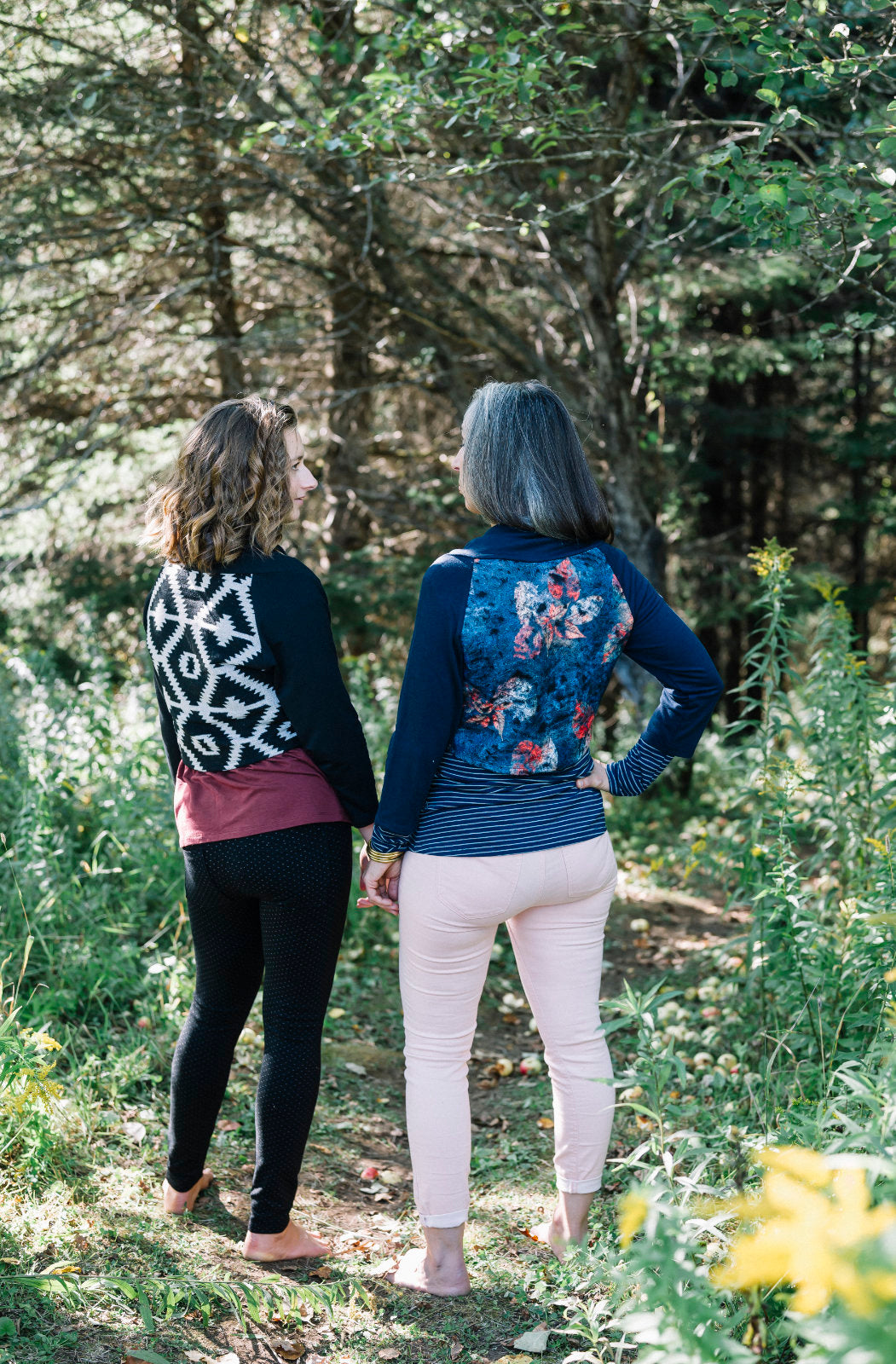 Back view of two women standing side to side in a forest, wearing the Boreal Bolero by Marie C. One is wearing the Black and one is wearing the Navy. The bolero features contrasting fabric at the back, a high collar that can be folded down, and a single button closure. 
