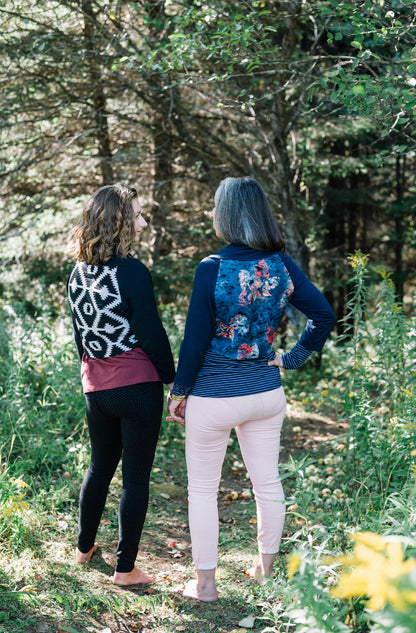 Back view of two women standing side to side in a forest, wearing the Boreal Bolero by Marie C. One is wearing the Black and one is wearing the Navy. The bolero features contrasting fabric at the back, a high collar that can be folded down, and a single button closure. 