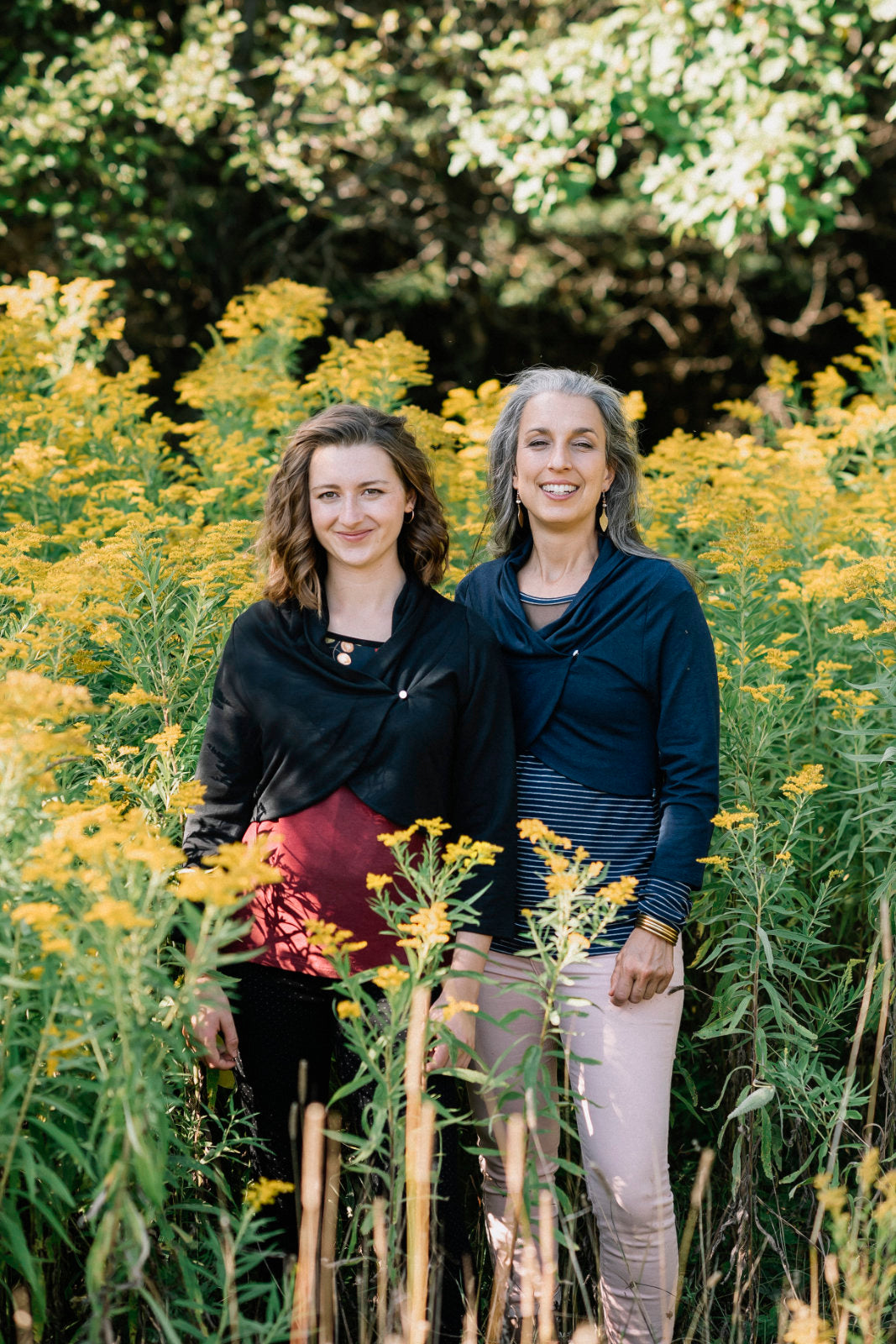 Two women standing side to side in a forest, wearing the Boreal Bolero by Marie C. One is wearing the Black and one is wearing the Navy. The bolero features contrasting fabric at the back, a high collar that can be folded down, and a single button closure. 