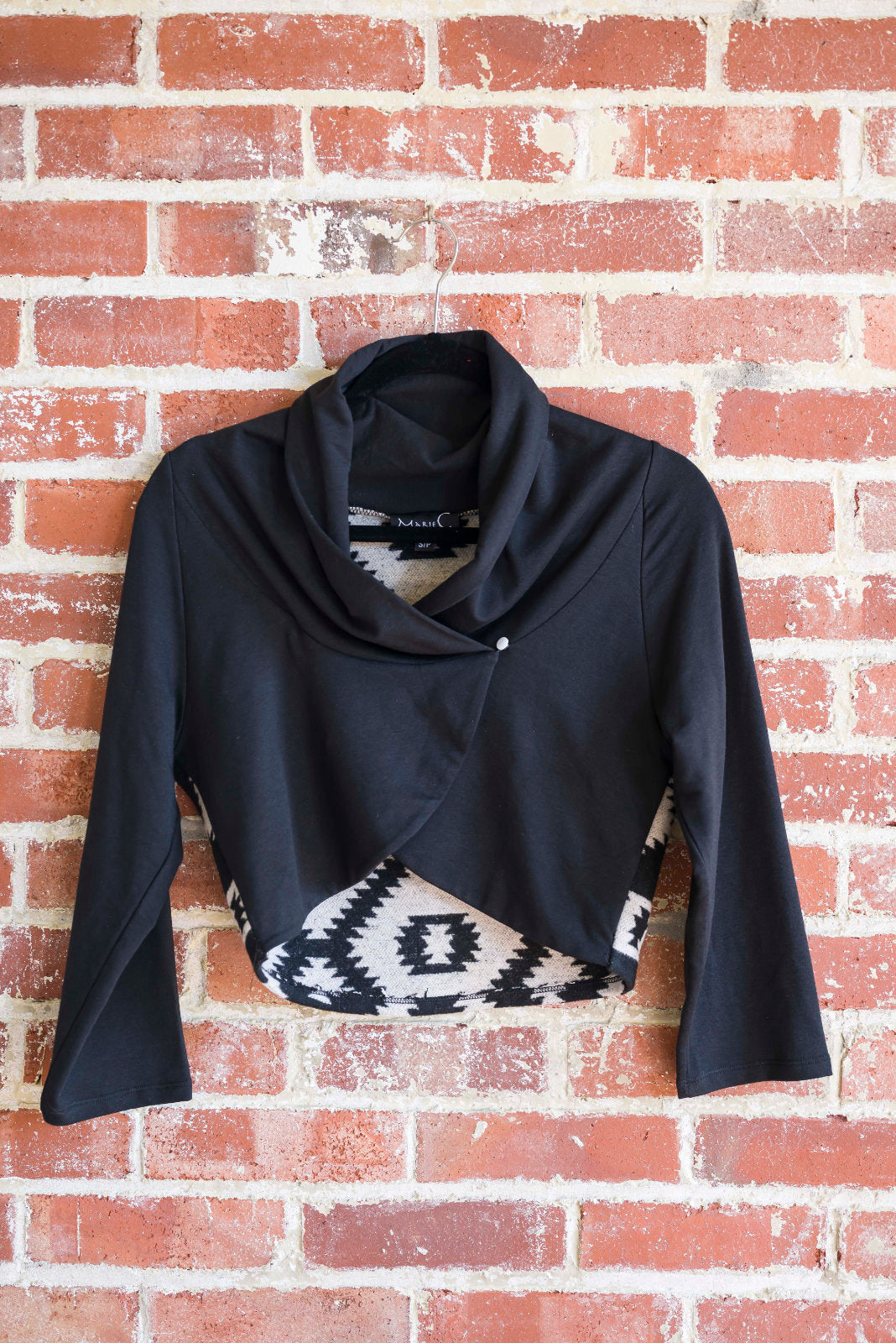 The Boreal Bolero by Marie C in Black is shown hanging against a brick wall. The bolero features contrasting fabric at the back, a high collar that can be folded down, and a single button closure. 