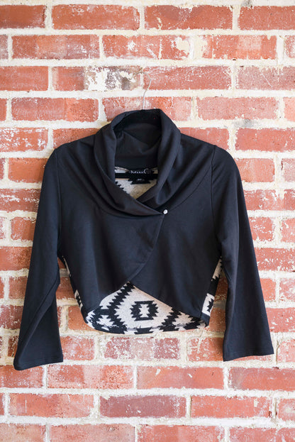 The Boreal Bolero by Marie C in Black is shown hanging against a brick wall. The bolero features contrasting fabric at the back, a high collar that can be folded down, and a single button closure. 