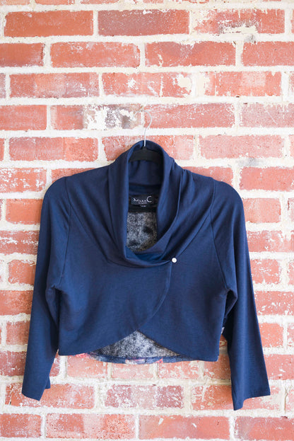 The Boreal Bolero by Marie C in Navy is shown hanging against a brick wall. The bolero features contrasting fabric at the back, a high collar that can be folded down, and a single button closure. 