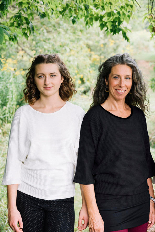 Two women wearing the Perla Top by Marie C,  one in Ivory and one in Black, with a wide round neck, 3/4 batwing sleeves, and fitted waist in ribbed knit fabric. They are standing outside. 
