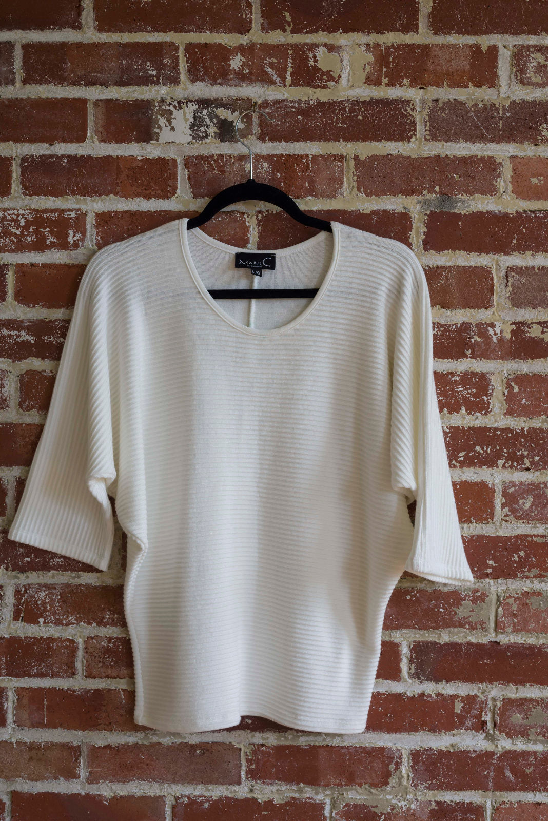 The Perla Top by Marie C in Ivory, with a wide round neck, 3/4 batwing sleeves, and fitted waist in ribbed knit fabric, is shown hanging against a brick wall. 
