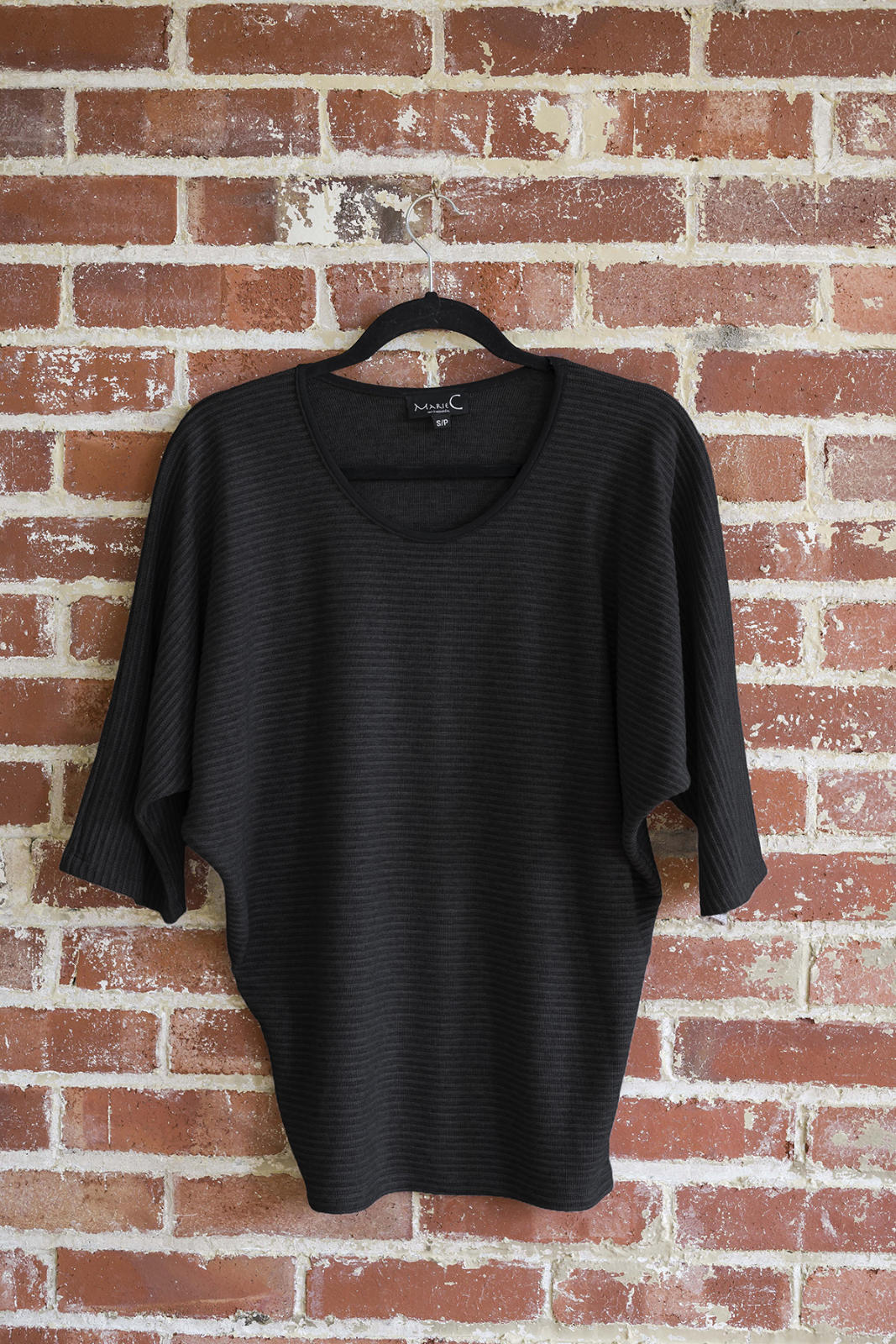 The Perla Top by Marie C in Black, with a wide round neck, 3/4 batwing sleeves, and fitted waist in ribbed knit fabric, is shown hanging against a brick wall. 