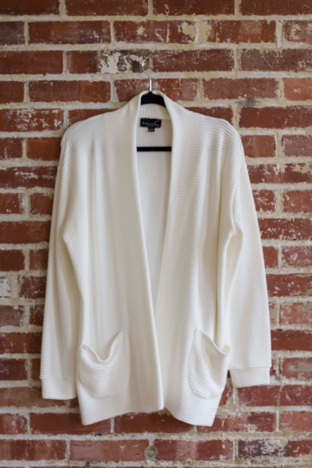 The Margo Cardigan by Marie C in Ivory, a long open cardigan  in ribbed knit with rounded patch pockets and dropped sleeves, is shown hanging in front of a brick wall. 