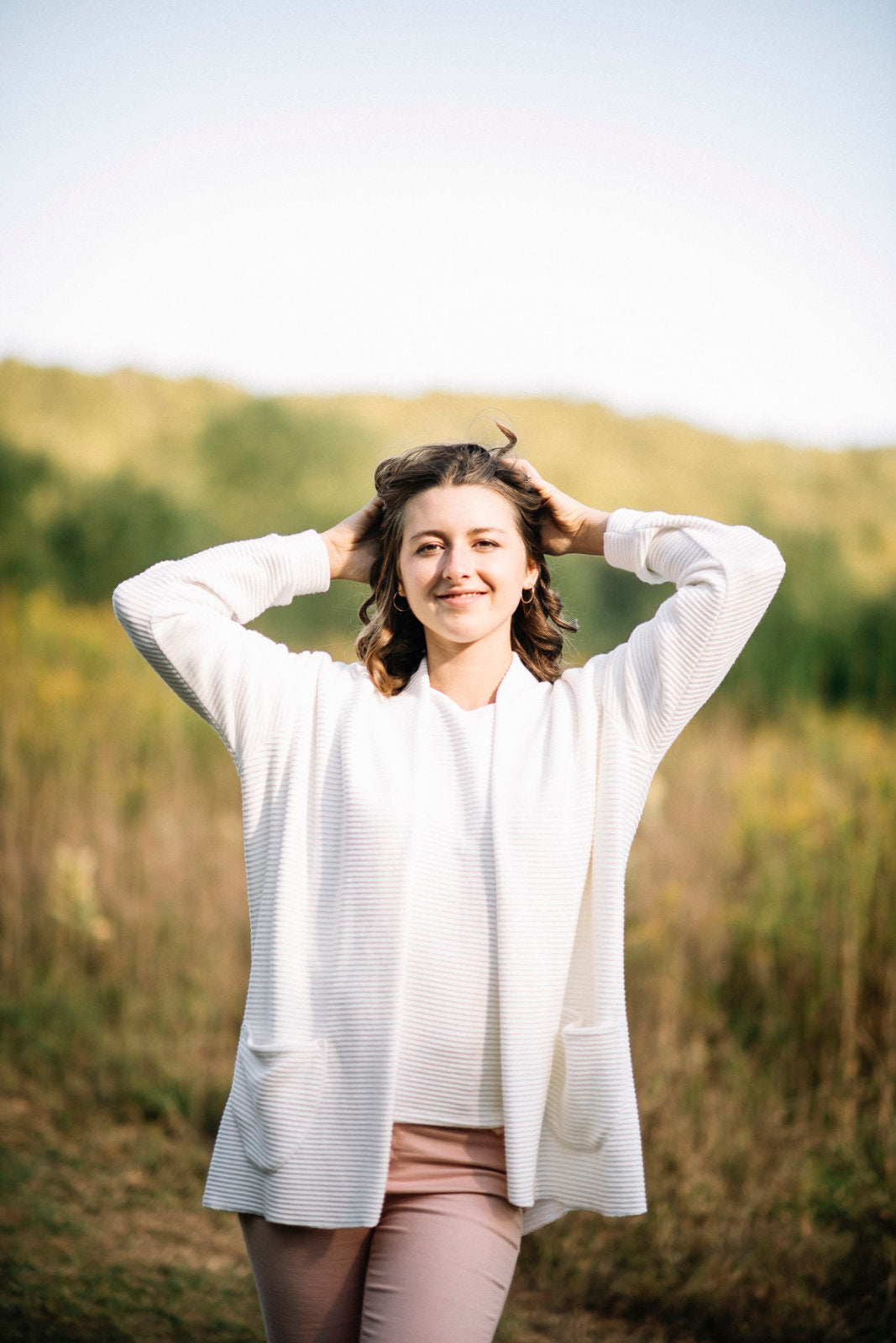 A woman wearing the Margo Cardigan by Marie C in Ivory, a long open cardigan  in ribbed knit with rounded patch pockets and dropped sleeves. She is wearing it with pink jeans and standing in a field. 
