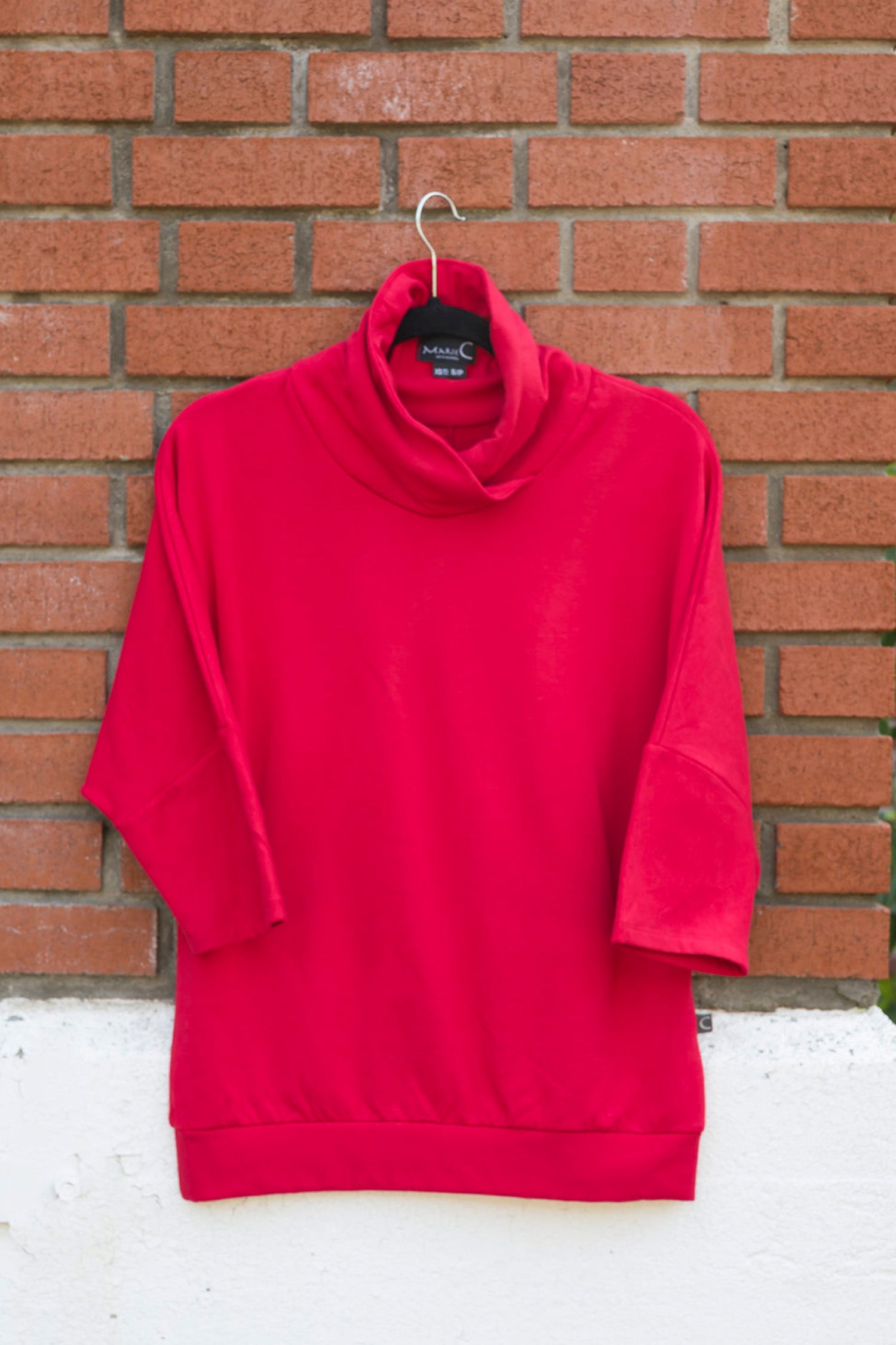 The Beaaa Top by Marie C in Red, hanging in front of a brick wall 