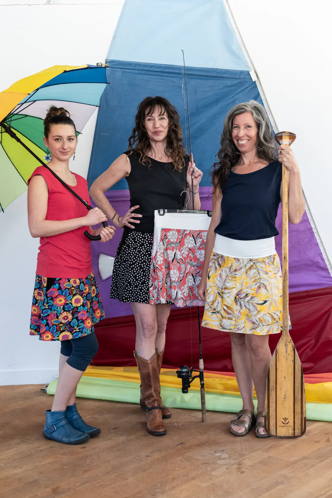 Three women wearing the Sol Top from Marie C, a reversible cap sleeve tee. From left to right they are wearing it in Coral, Black and Navy. They are all wearing skirts and are standing inside in front of a sail with outdoor accessories. 
