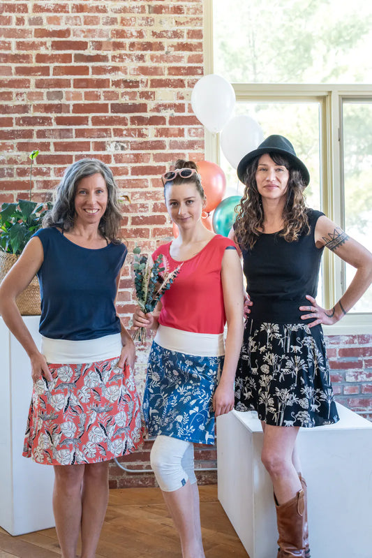 Three women wearing the Sol Top from Marie C, a reversible cap sleeve tee. From left to right they are wearing it in Navy, Coral, and Black. They are all wearing skirts and are standing at a party. 