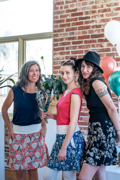Three women wearing the Sol Top from Marie C, a reversible cap sleeve tee. From left to right they are wearing it in Navy, Coral, and Black. They are all wearing skirts and are standing at a party. 