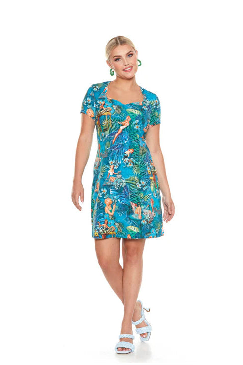 A woman wearing the Marilyn Dress by Yul Voy, featuring a vintage inspired tropical print with pin-up girls on it, a sweetheart neckline, short sleeves, and a sheath shape that falls above the knee. 