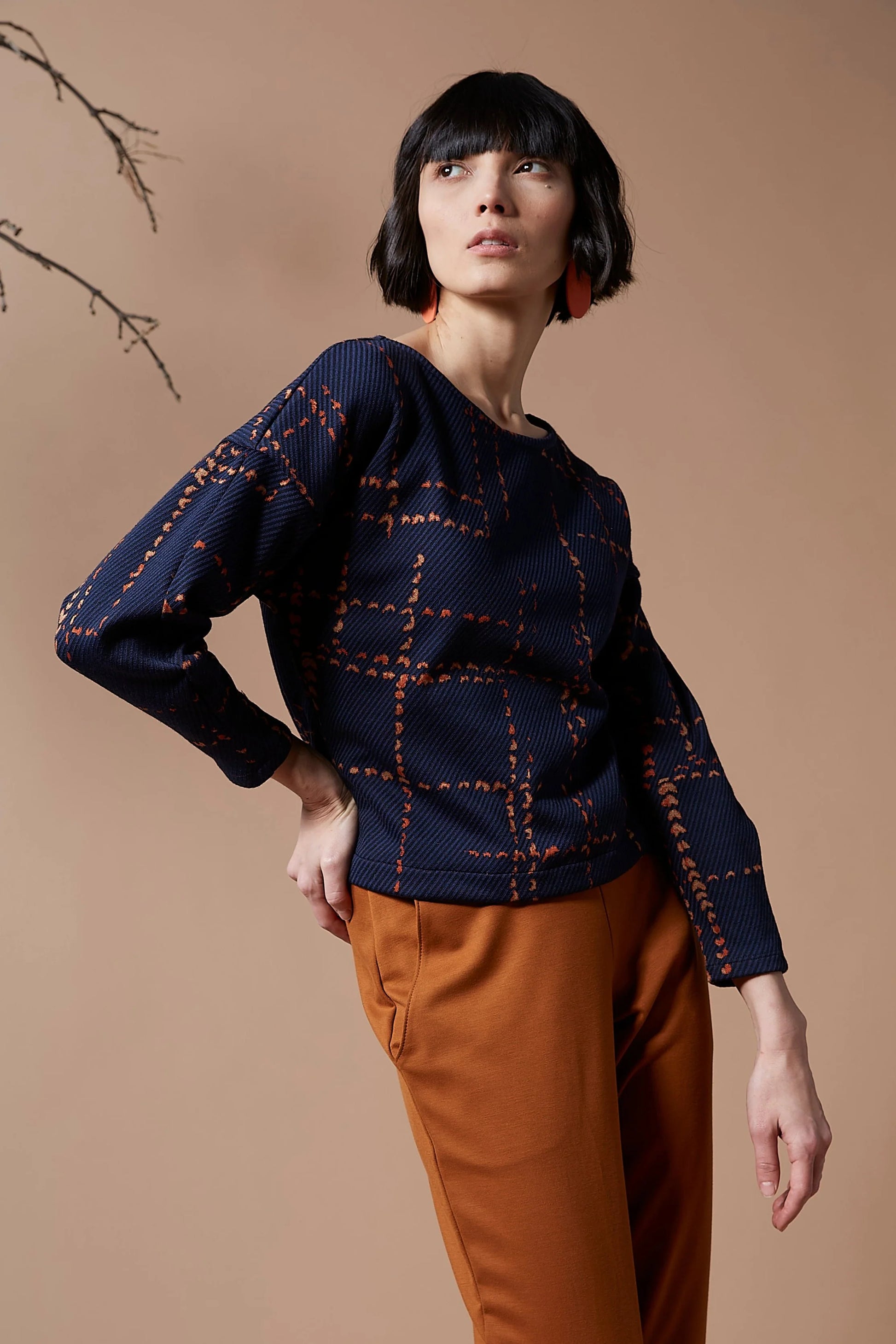 A woman wearing the Jackson Top by Melow in indigo with orange pants, standing in front of a peach background 