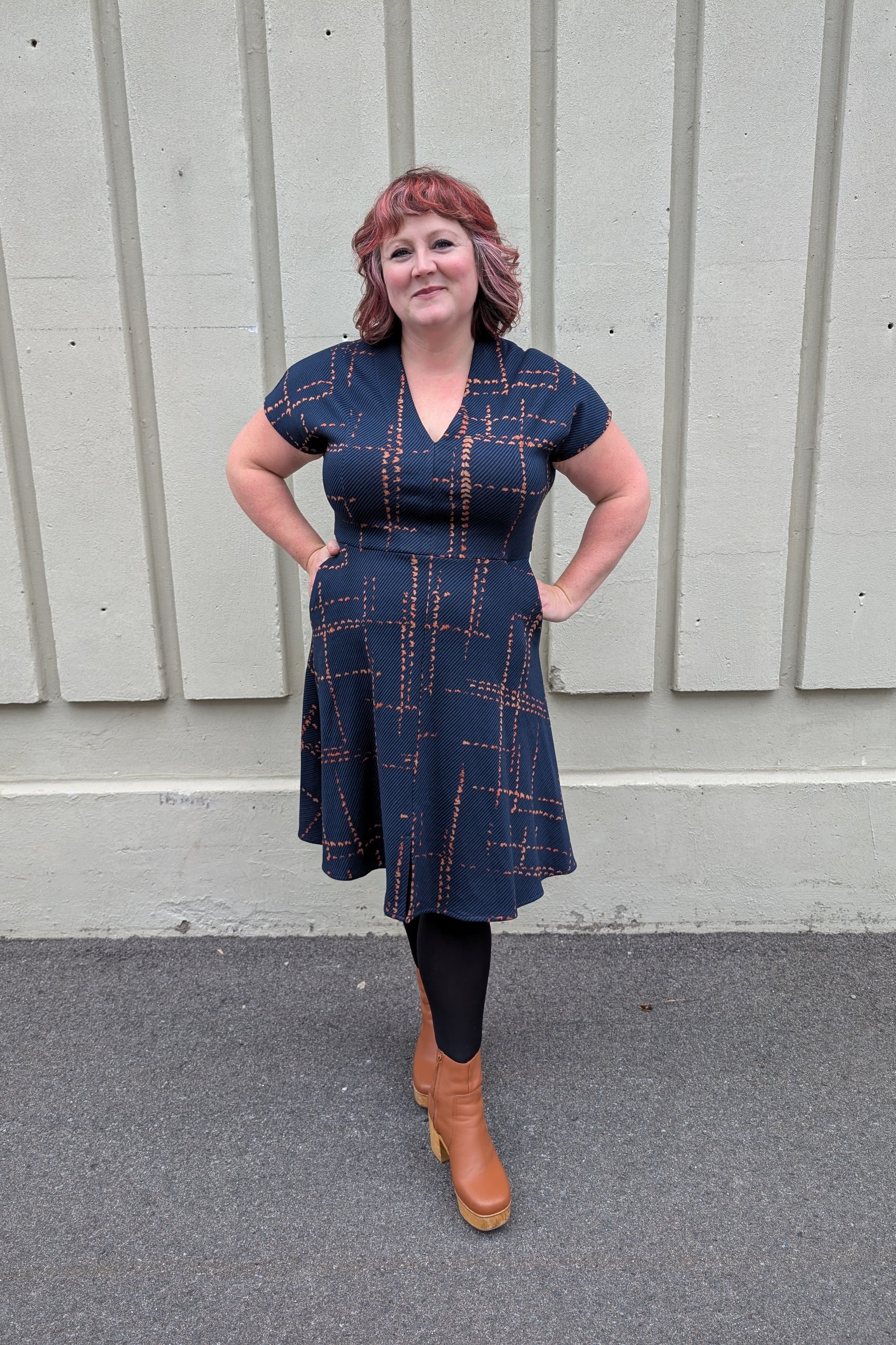 A woman wearing the Josee Dress by Melow in Indigo with orange plaid, featuring a V-neck, short sleeves, a defined waist, a fit and flare shape, and as slit at the front of the knee-length hem. 