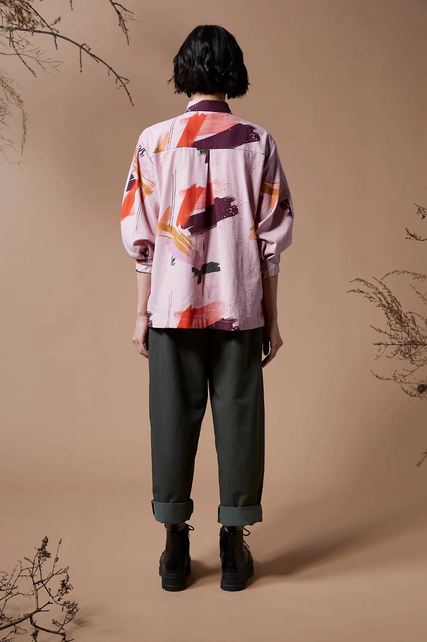 Back view of a woman wearing the Juliette Top by Melow in Sobel Print, featuring a pale pink background with splashes of colour.  The shirt has a blouse collar, button front, and 3/4 length puff sleeves. 