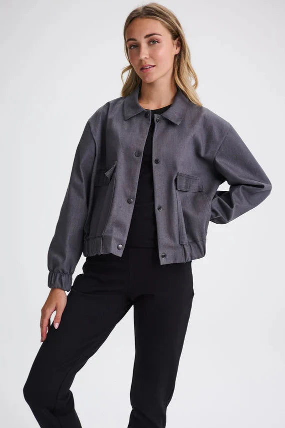 A woman wearing the Charlevoix Denim Bomber Jacket by Message Factory in Grey, with a shirt collar, button front, and large patch pockets, with black pants, standing in front of a white background 