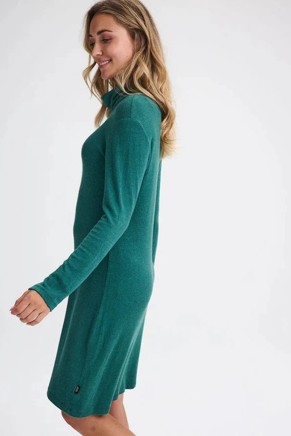 Side view of a woman wearing the Betula Dress by Message Factory in Evergreen, a knit turtleneck dress with long sleeves and a knee-length hem. She is standing in front of a beige background. 