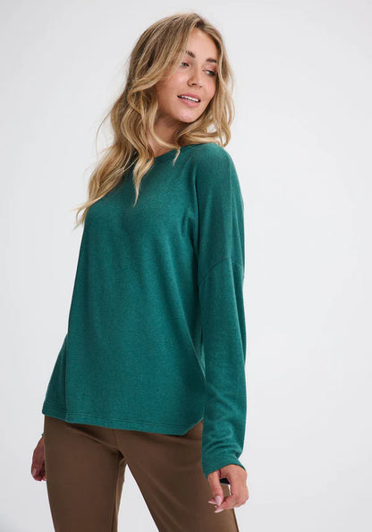 A woman wearing the Chandler Top by Message Factory in Green, with a wide neck, drop shoulders, long sleeves, and a hi-low, made from bamboo and cotton, with brown pants. She's standing in front of a white background. 