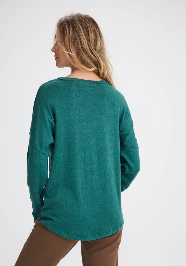 Back view of a woman wearing the Chandler Top by Message Factory in Green, with a wide neck, drop shoulders, long sleeves, and a hi-low, made from bamboo and cotton, with brown pants. She's standing in front of a white background. 