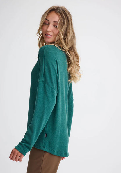 Side view of a woman wearing the Chandler Top by Message Factory in Green, with a wide neck, drop shoulders, long sleeves, and a hi-low, made from bamboo and cotton, with brown pants. She's standing in front of a white background. 