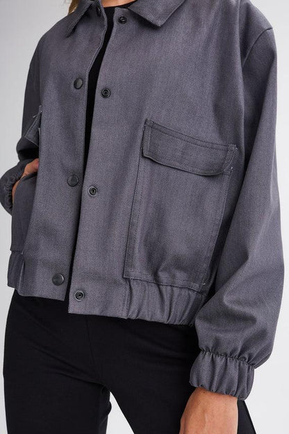 Neck down close-up of a woman wearing the Charlevoix Denim Bomber Jacket by Message Factory in Grey, with a shirt collar, button front, and large patch pockets, with black pants, standing in front of a white background 