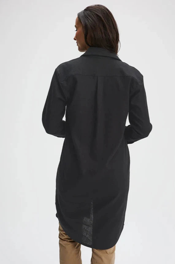 Back view of a woman wearing the Donnaconna Shirt Dress by Message Factory in Black, with a button front, classic collar, and rounded hem, made from natural ramie fabric. She is wearing it with beige pants and is standing in front of a white background. 