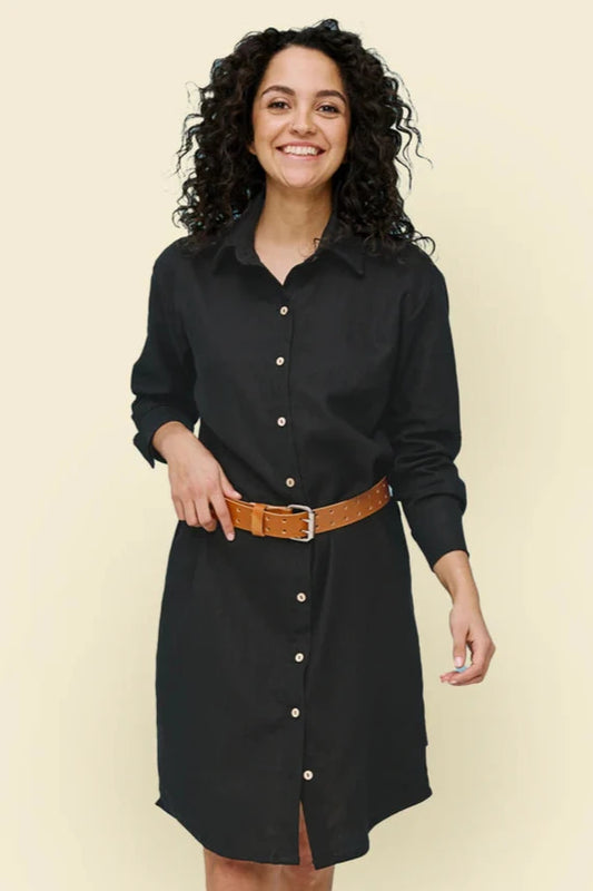 A woman wearing the Donnaconna Shirt Dress by Message Factory in Black, with a button front, classic collar, and rounded hem, made from natural ramie fabric. She is wearing it with a tan belt and is standing in front of a white background. 