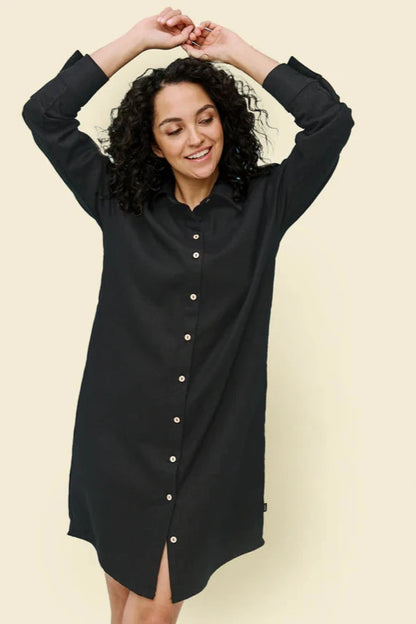 A woman wearing the Donnaconna Shirt Dress by Message Factory in Black, with a button front, classic collar, and rounded hem, made from natural ramie fabric. She is standing in front of a white background. 