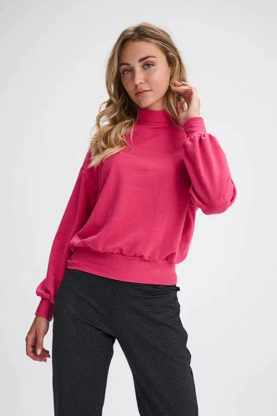 A woman wearing the Fermont Sweatshirt by Message Factory in Fuchsia, made from bamboo and cotton with a high neck, long sleeves with a puff detail at the cuffs, and fitted band at the bottom. She is wearing it with grey pants and is standing in front of a white background 