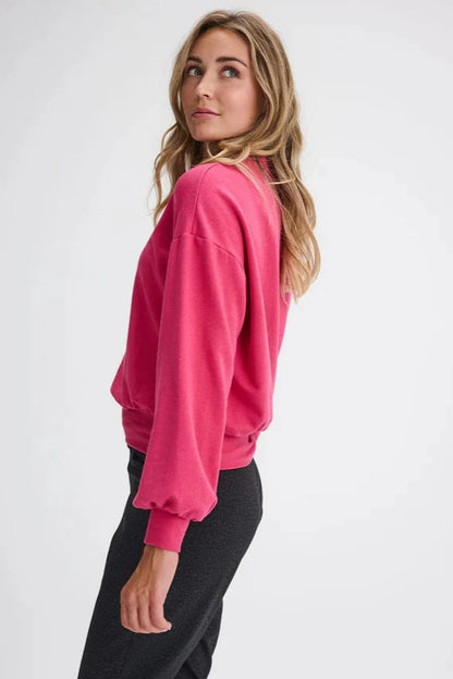 Side view of a woman wearing the Fermont Sweatshirt by Message Factory in Fuchsia, made from bamboo and cotton with a high neck, long sleeves with a puff detail at the cuffs, and fitted band at the bottom. She is wearing it with grey pants and is standing in front of a white background. 