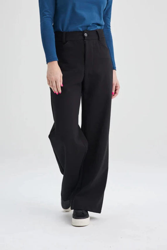 A waist down view of a woman wearing the Hatley Pants by Message Factory in Black, a wide leg pant with a button fly and two pockets, made from Lenzing Ecovero viscose, with a blue top. She is standing in front of a white background. 