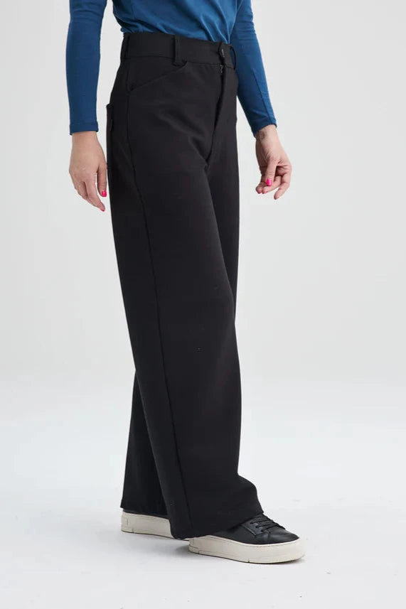 A waist down view of a woman wearing the Hatley Pants by Message Factory in Black, a wide leg pant with a button fly and two pockets, made from Lenzing Ecovero viscose, with a blue top. She is standing in front of a white background. 